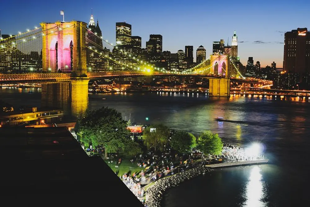 The image shows a vibrant night view of the Brooklyn Bridge illuminated in pink and yellow lights with the Manhattan skyline in the background and a bustling waterfront promenade along the East River