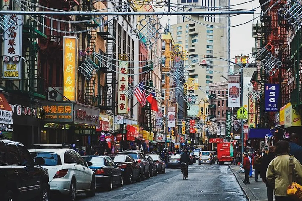 A bustling urban street is lined with colorful signs in various languages decorated with festive lanterns and filled with pedestrians and parked cars conveying a vibrant Chinatown atmosphere