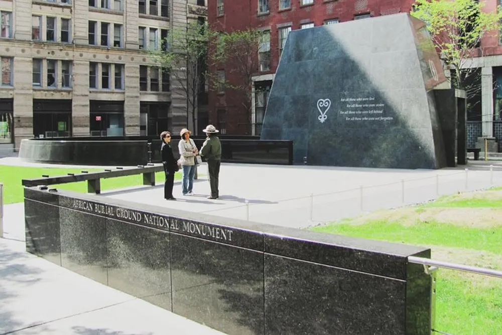 Three individuals are having a conversation next to a placard that reads African Burial Ground National Monument with a large memorial structure in the background among city buildings