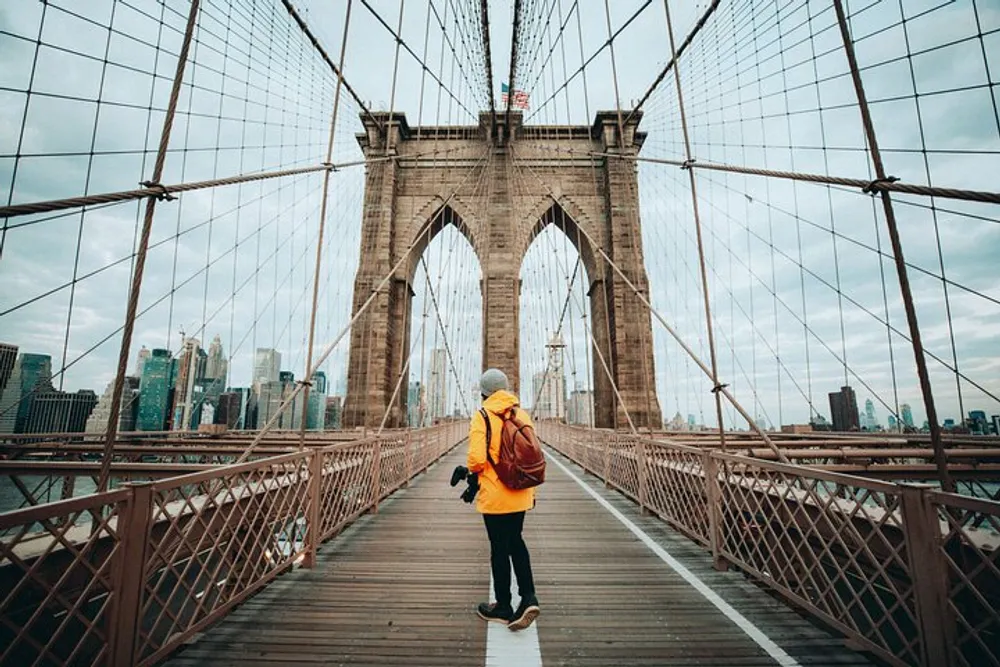 A person wearing a yellow jacket and carrying a backpack and camera is walking across the Brooklyn Bridge with the New York City skyline in the background