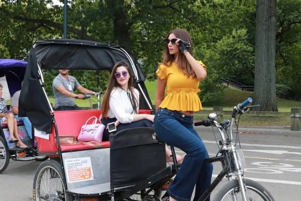 Two women one seated in a pedicab and the other standing beside it are both wearing sunglasses and appear to be posing for a photo in a park setting