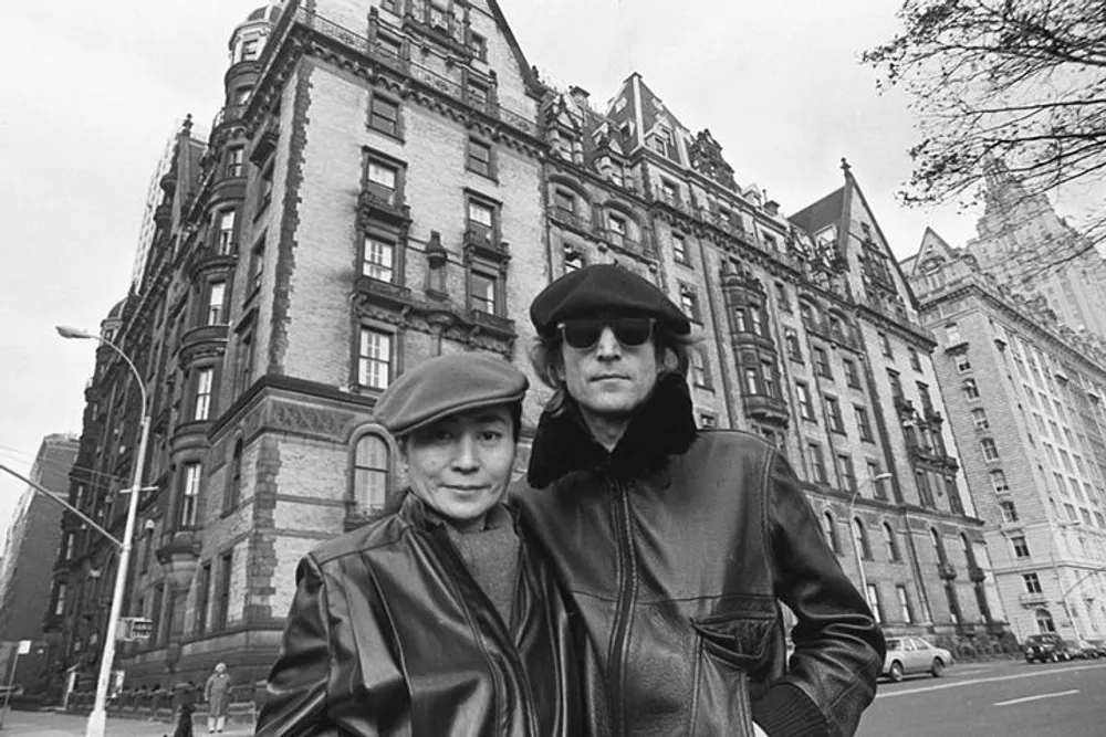 Two people stand in front of a grand old building both dressed in leather jackets and one wearing a beret the other a cap seeming to exude a cool and artistic vibe