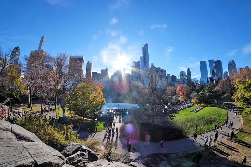The image depicts a vibrant and sunny day in Central Park with visitors enjoying the natural scenery framed by the towering skyscrapers of Manhattan in the background
