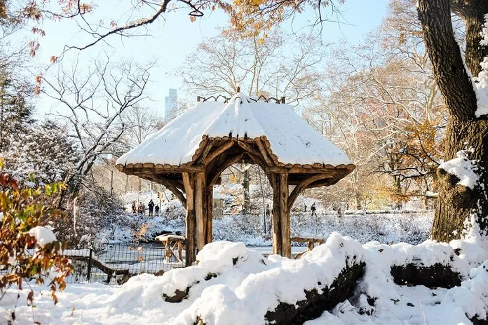 A wooden gazebo topped with a layer of fresh snow stands in a tranquil snow-covered park with people in the distance and skyscrapers peeking through bare trees under a clear sky