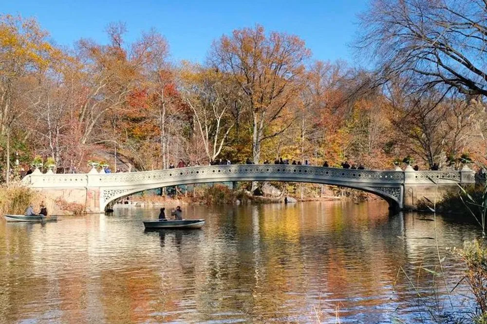 The image depicts a scenic view of a bridge over a tranquil lake with people rowing boats and others walking on the bridge surrounded by autumn-colored trees under a clear blue sky