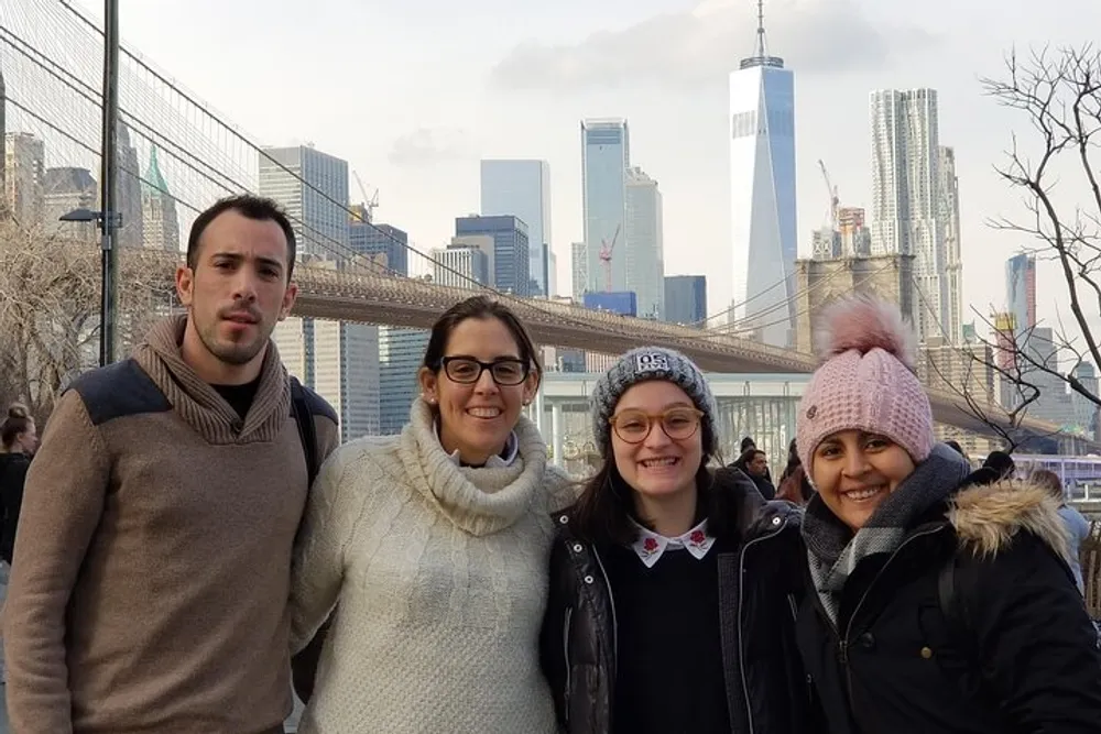 Four people are posing for a photo with the Brooklyn Bridge and the New York City skyline including the One World Trade Center in the background