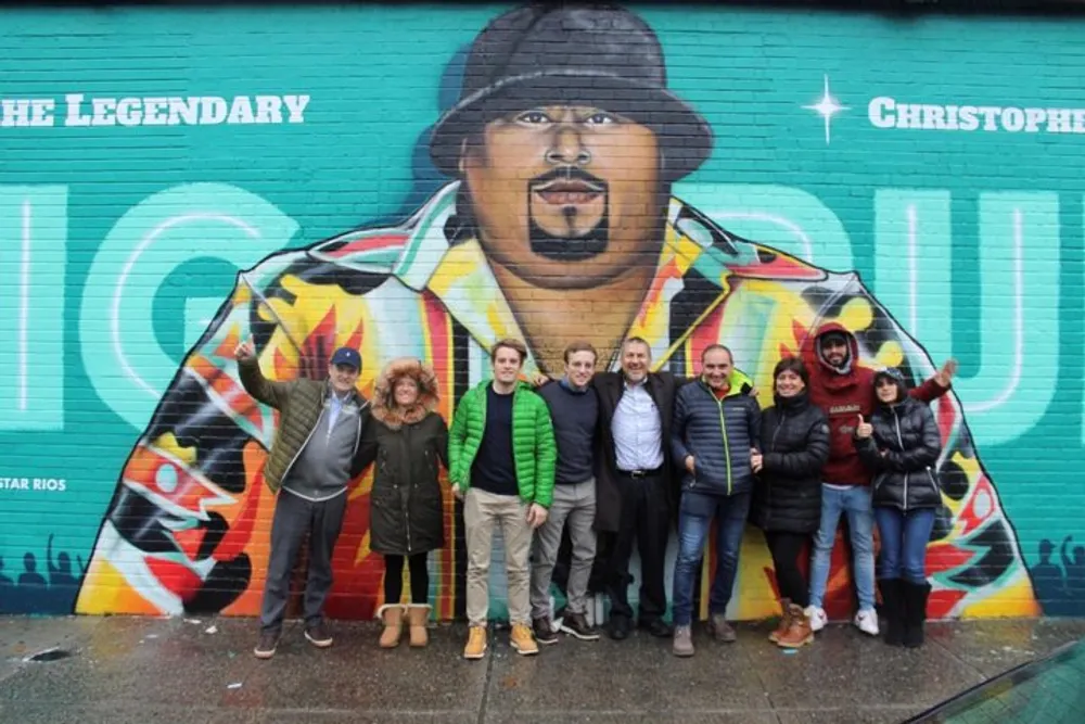 A group of people is posing for a photo in front of a colorful mural of a man with the text The Legendary Christopher BIG Wallace behind them