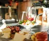 Red wine is being poured into a glass with a cheese platter in the foreground and a blurred background suggesting a cozy dining atmosphere