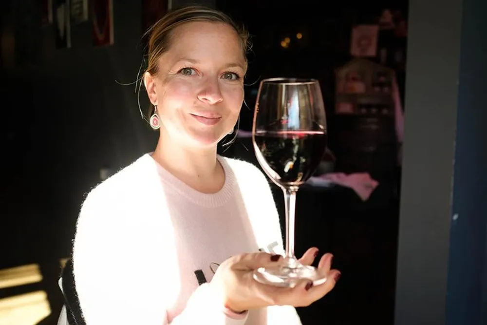 A smiling woman is holding a glass of red wine in a room with sunlight streaming in