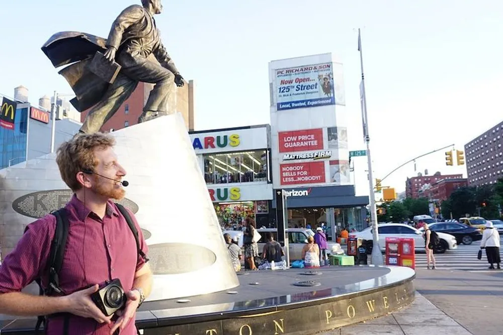 A man holding a camera stands in front of a statue of a historical figure in an urban square with commercial signs in the background