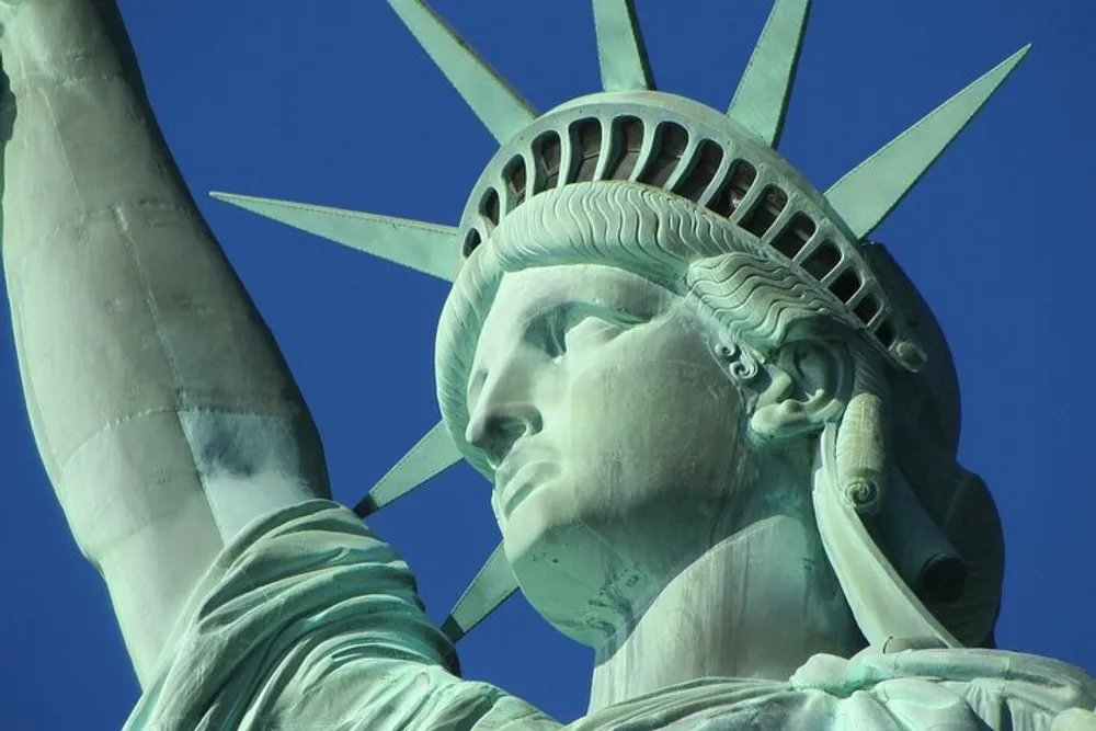 The image shows a close-up of the Statue of Libertys head and raised torch against a clear blue sky