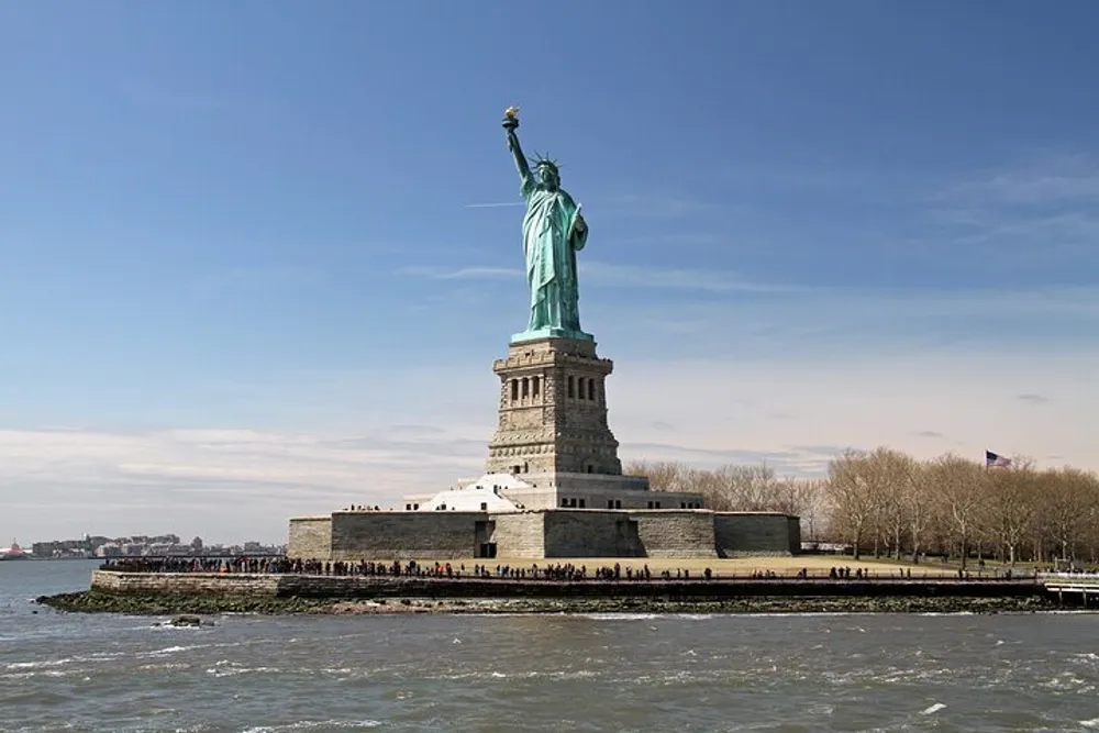 The Statue of Liberty stands tall against a clear sky on Liberty Island surrounded by water and visitors