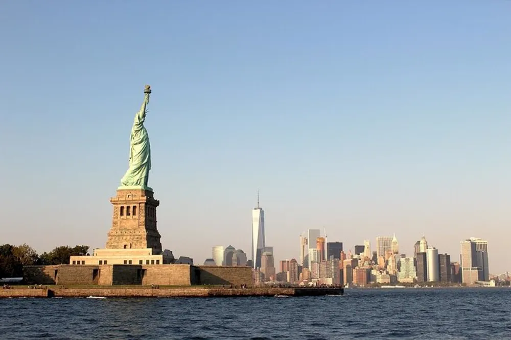 The Statue of Liberty stands prominently in the foreground with the New York City skyline including the One World Trade Center in the background