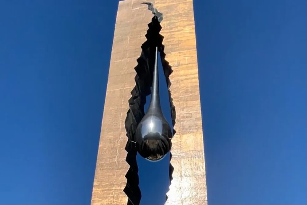 The image shows a view from below of a towering narrow sculpture with a metallic reflective surface and a jagged central split against a clear blue sky
