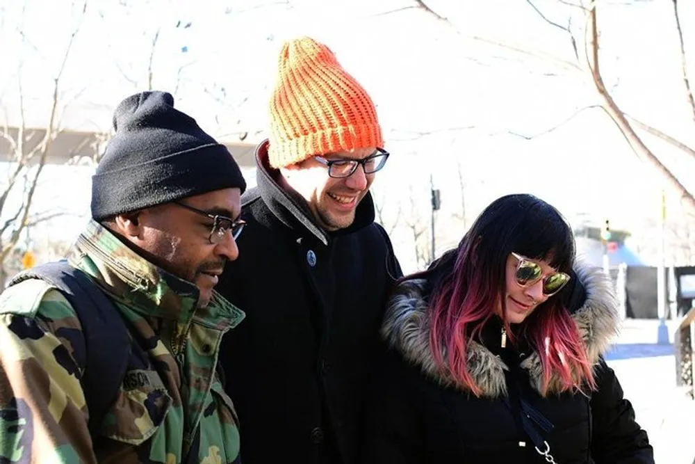 Three people are smiling and looking downward possibly at something interesting out of frame while dressed in warm clothes suggesting a cool outdoor setting