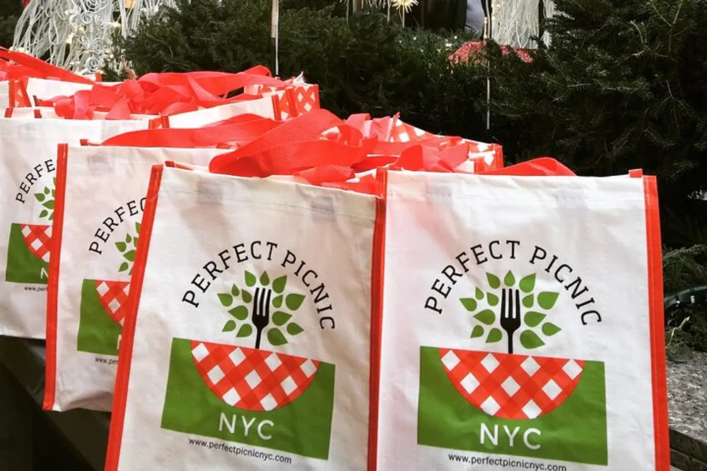 The image shows a collection of white tote bags with red handles and Perfect Picnic NYC branding set against a backdrop of greenery