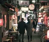 A couple is walking down a vibrant alley illuminated by neon signs and traditional lanterns evoking the bustling atmosphere of an urban Japanese dining district