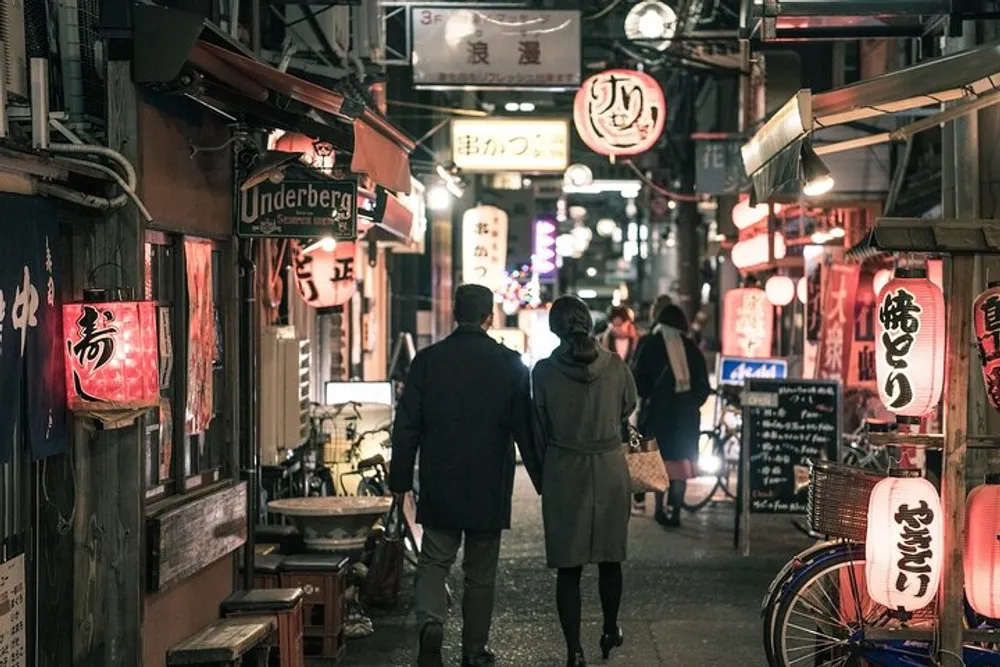 A couple is walking down a vibrant alley illuminated by neon signs and traditional lanterns evoking the bustling atmosphere of an urban Japanese dining district