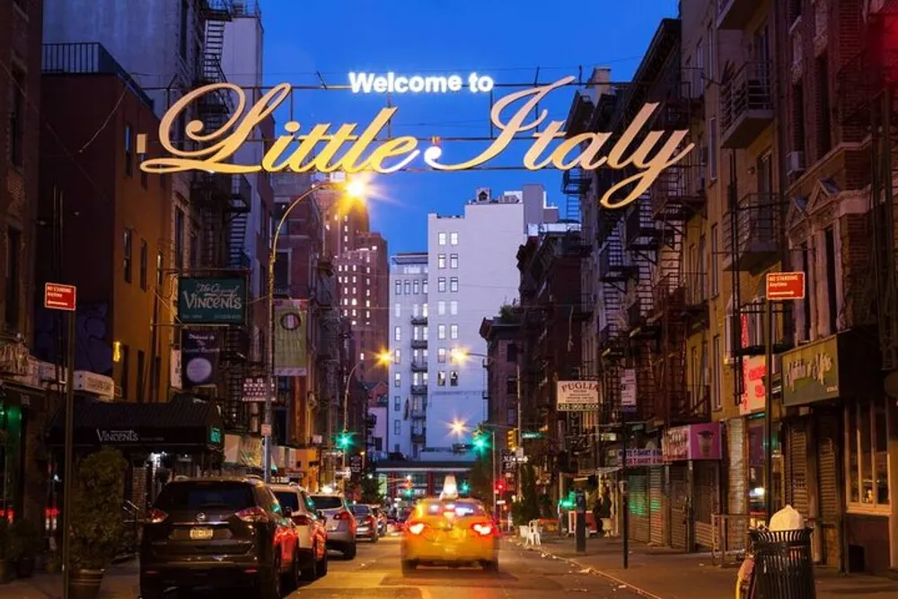 The image shows a bustling street scene at twilight with the illuminated sign Welcome to Little Italy arching over the road indicating the entrance to a neighborhood known for its Italian heritage and cuisine