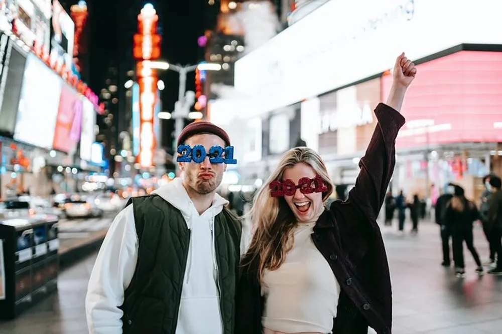 Two people are posing for a photo wearing 2024-themed novelty glasses at night with a brightly lit city street behind them