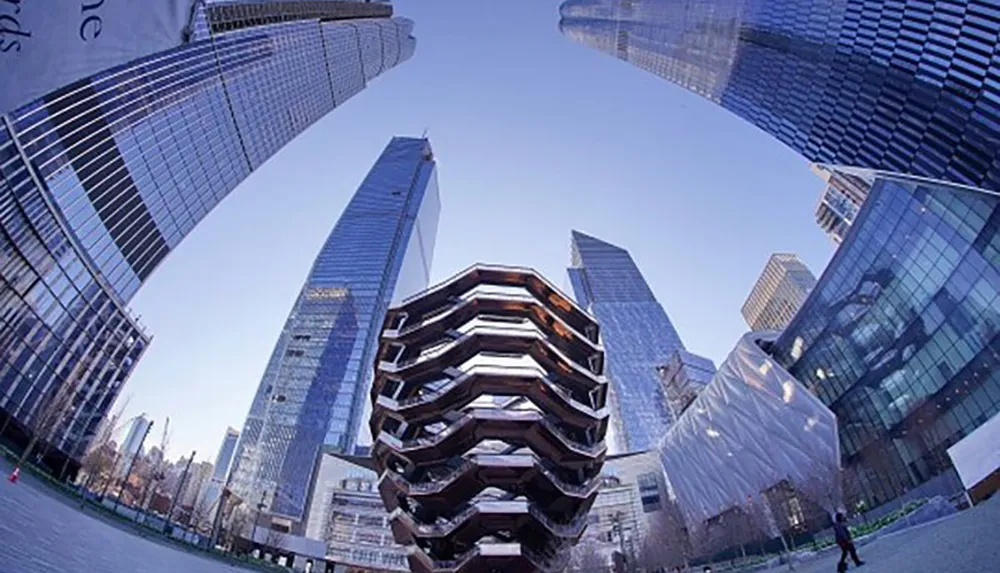 The image shows a person walking near the Vessel sculpture surrounded by towering skyscrapers of Hudson Yards in New York City