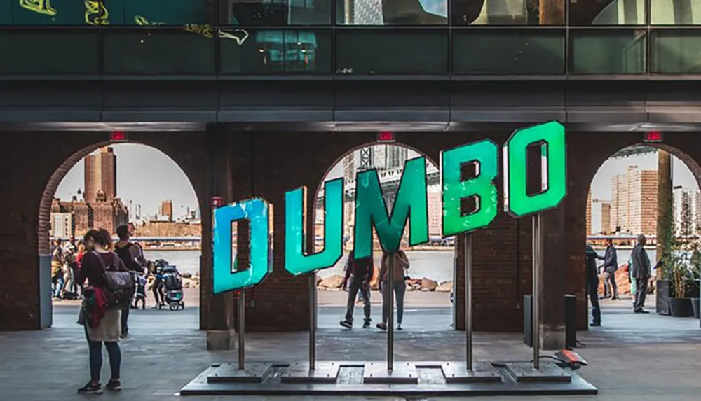 A large colorful sign spelling DUMBO is prominently displayed in an urban setting with people walking around it and a cityscape in the background