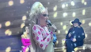 A performer with elaborate makeup and a large blonde hairstyle is speaking into a microphone on a stage with audience members looking on.