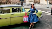 A smiling woman in a blue vintage-style dress is standing next to a classic green and yellow car while holding a shopping bag with city streets in the background.