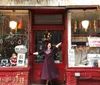 A person in a clown-like outfit is cheerfully posing with arms outstretched in front of an old-fashioned red storefront adorned with holiday decorations and a sign for Albanese Meats  Poultry