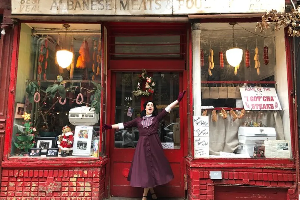 A person in a clown-like outfit is cheerfully posing with arms outstretched in front of an old-fashioned red storefront adorned with holiday decorations and a sign for Albanese Meats  Poultry
