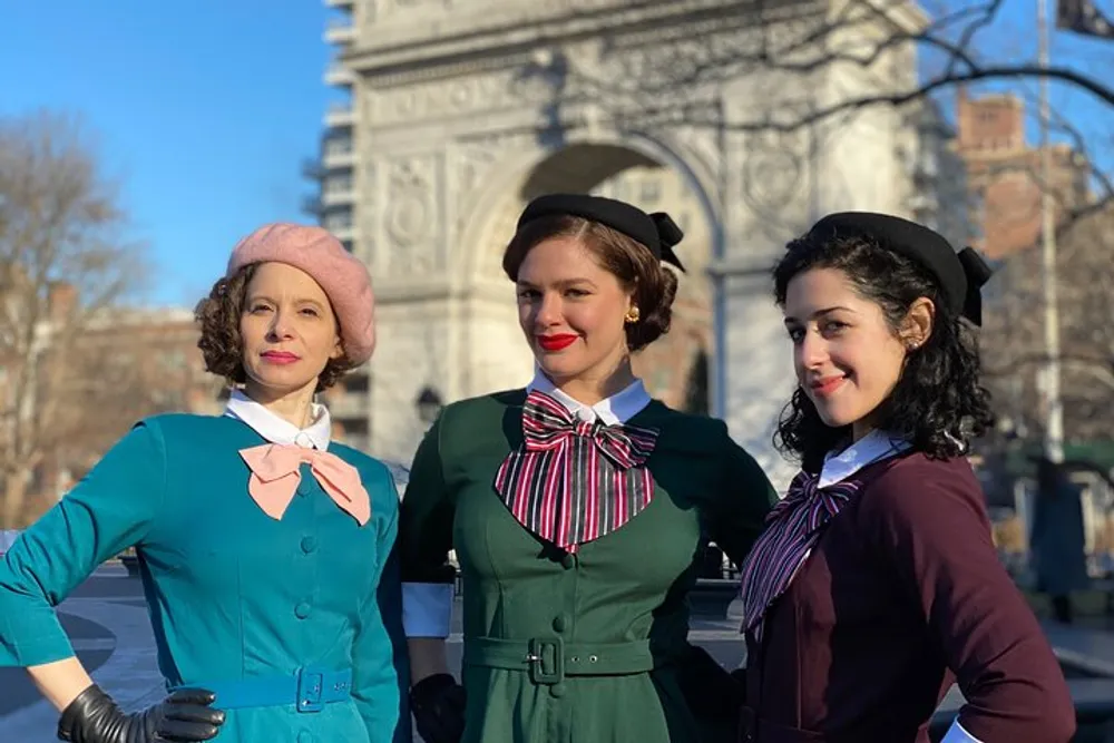 Three women are dressed in vintage-style outfits reminiscent of the 1940s or 1950s posing confidently with a historical arch structure in the background