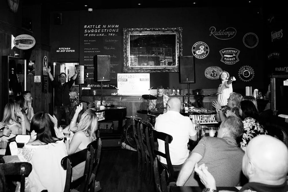 A black and white photo capturing a lively scene inside a pub with people seated at tables one person standing with a raised hand and a small stage where another person is speaking into a microphone