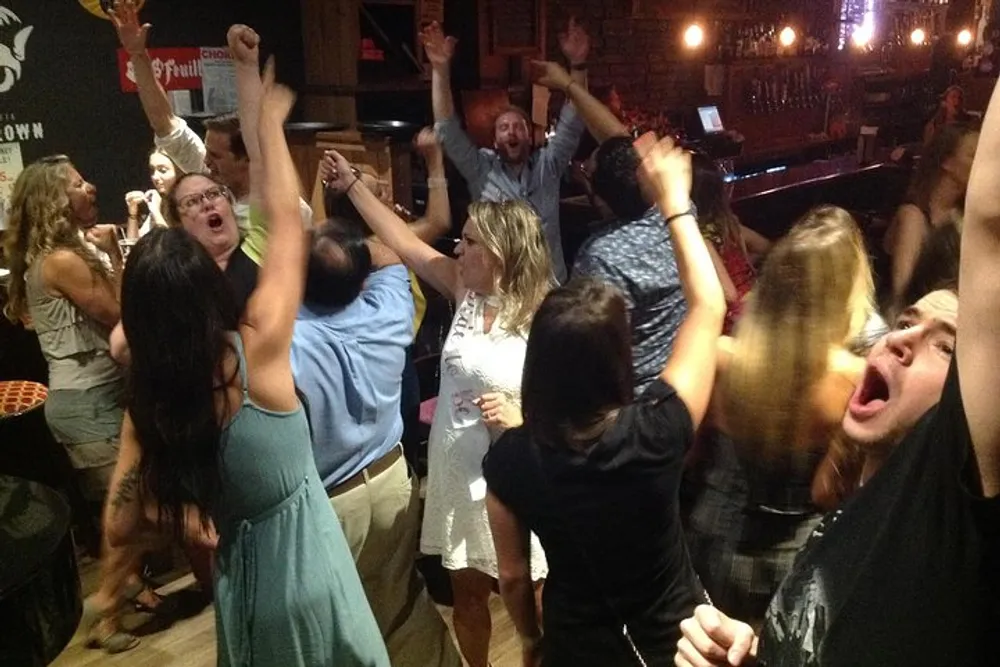 A group of people is enthusiastically dancing and celebrating some with their arms raised in what appears to be a lively indoor party environment