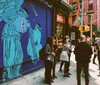 A group of people are standing on a city street corner in front of a striking blue building mural that features an angelic figure