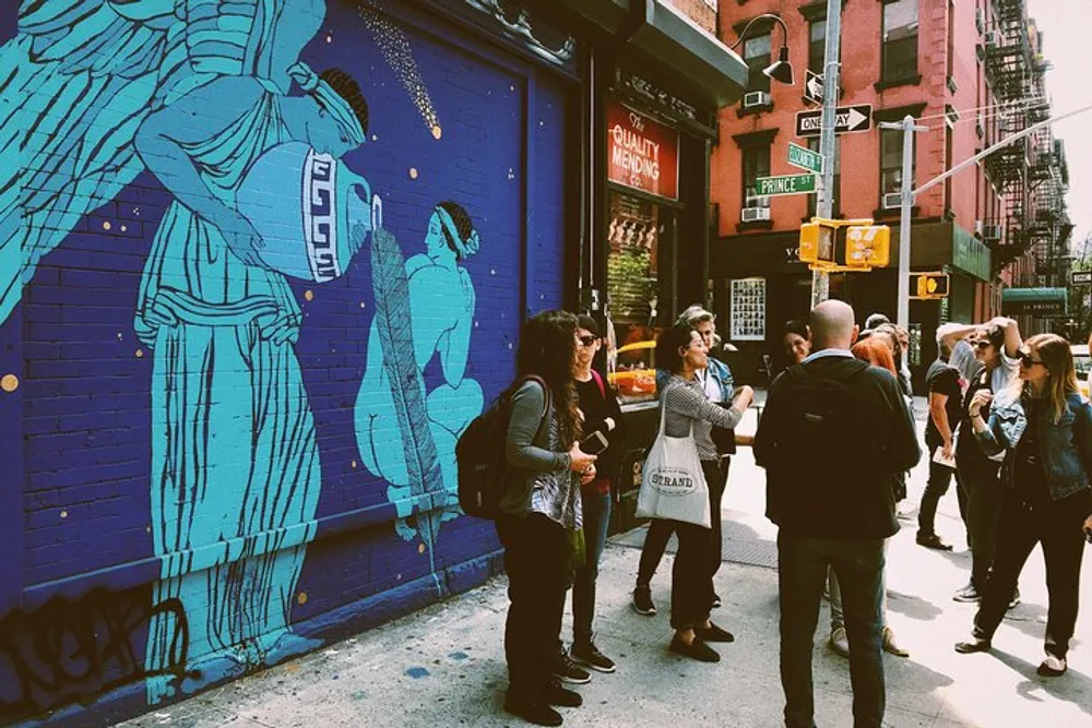 A group of people are standing on a city street corner in front of a striking blue building mural that features an angelic figure