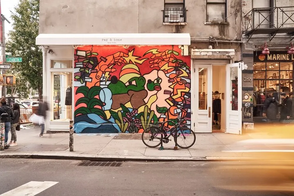 A vibrant street mural adorns the exterior wall of a building on a bustling city street with passersby and a parked bicycle in the foreground