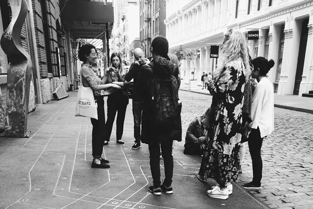 A group of people are engaged in conversation on a city street with hopscotch markings on the pavement