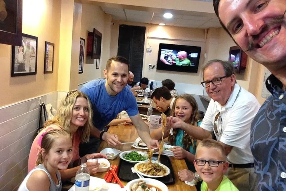 A group of people is smiling at the camera while enjoying a meal together at a restaurant with a TV in the background