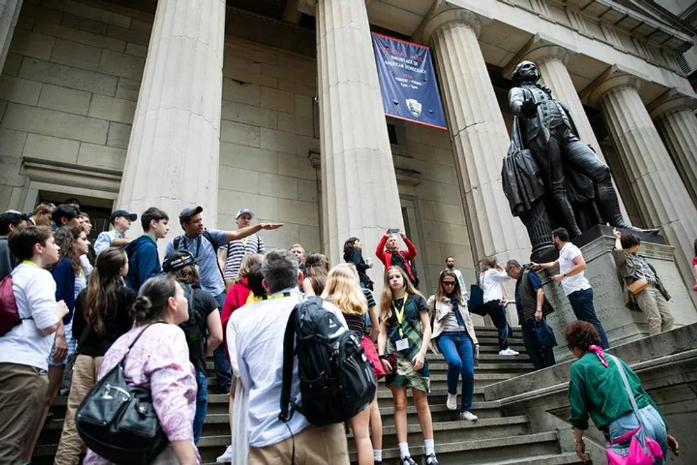 A group of people possibly on a tour are gathered on the steps of a building next to a large statue with one person pointing out something of interest