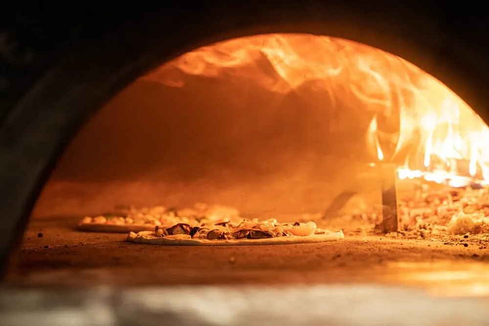 A wood-fired pizza oven is baking pizza showing the glowing flames and the pizza cooking on the oven floor