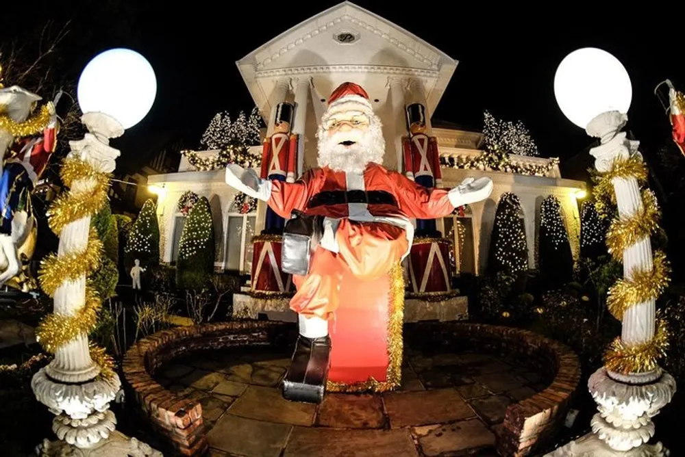 A large inflatable Santa Claus decoration stands in front of a festively decorated house with illuminated trees and holiday adornments at nighttime