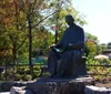 The image shows a bronze statue of a seated figure reading a book situated in a park-like setting with trees and a clear sky in the background