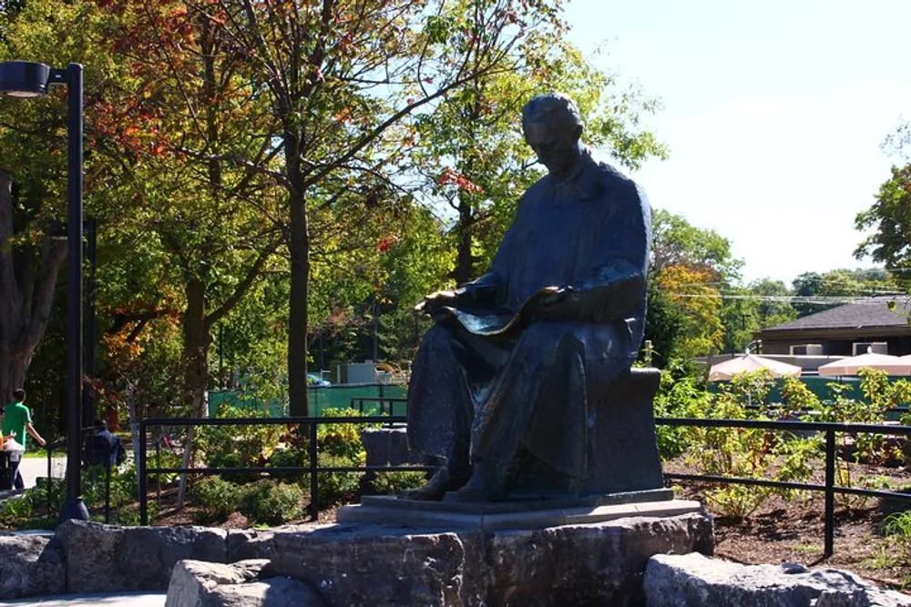 The image shows a bronze statue of a seated figure reading a book situated in a park-like setting with trees and a clear sky in the background