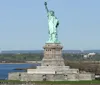 The image shows the Statue of Liberty standing tall on Liberty Island with a clear blue sky in the background and visitors gathered around its base