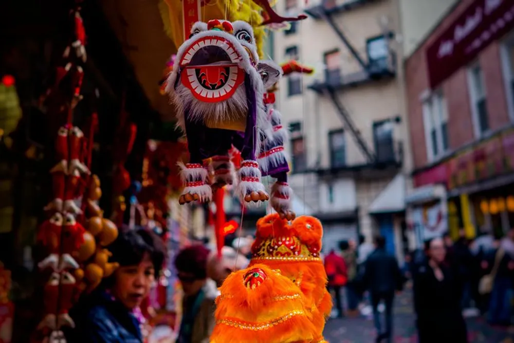 A colorful Chinese lion dance costume is hanging among other festive decorations in a bustling street market scene with a blurred view of onlookers in the background