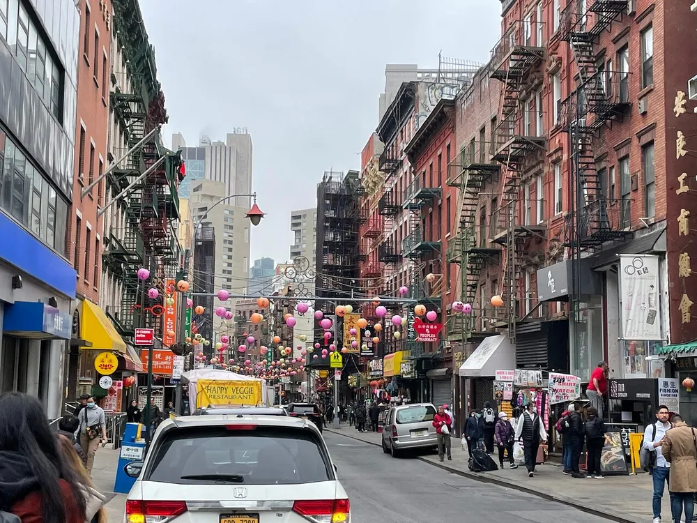 The image shows a vibrant street scene likely in a Chinatown district with traditional lanterns hanging above numerous signs in Chinese charming old buildings with fire escapes and pedestrians walking along the sidewalk