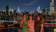 Three smiling women pose together at night with a vibrant city skyline illuminated in the background.