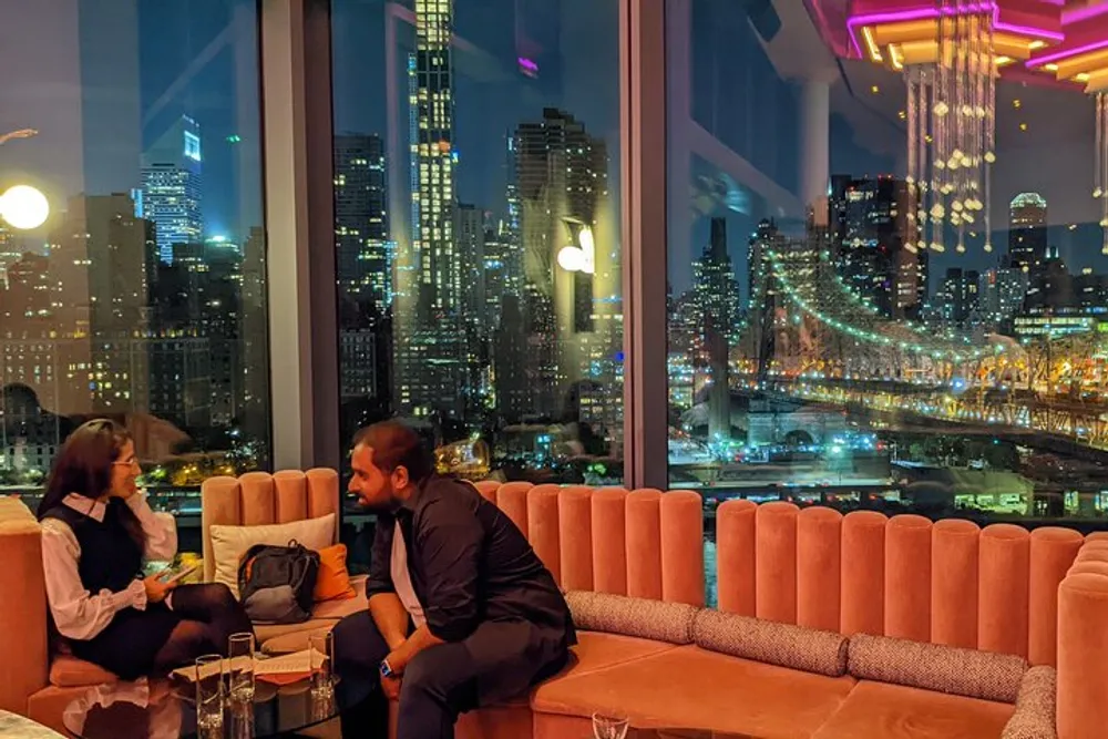 Two people are engaged in a conversation on a plush sofa with a vibrant night view of a cityscape and lit-up bridge seen through the expansive windows behind them