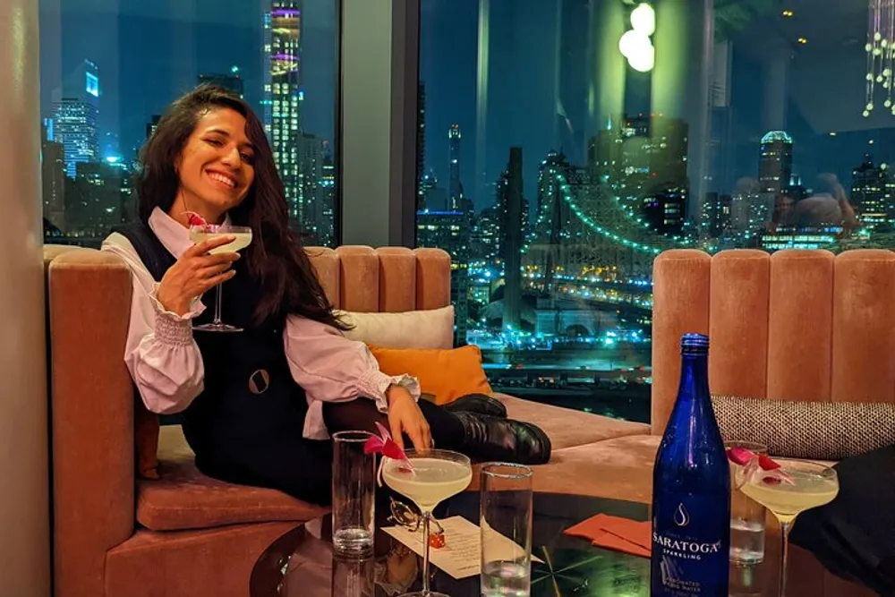 A person is smiling and holding a cocktail at a lounge with a vibrant city nightscape visible through the window behind them