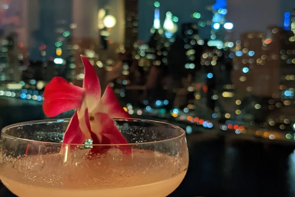 A cocktail garnished with a red flower sits in the foreground with a blurred nightscape of city lights in the background